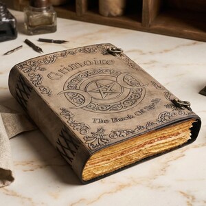 K&ouml;nnte beinhalten: Ein ledergebundenes Buch mit den Worten "Grimoire" und "The Book of Spells" auf dem Einband. Der Einband zeigt ein Pentagramm und ein Monddesign. Das Buch hat gealterte, vergilbte Seiten und Metallverschl&uuml;sse.