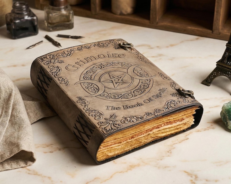 K&ouml;nnte beinhalten: Ein ledergebundenes Buch mit den Worten "Grimoire" und "The Book of Spells" auf dem Einband. Der Einband zeigt ein Pentagramm und ein Monddesign. Das Buch hat gealterte, vergilbte Seiten und Metallverschl&uuml;sse.