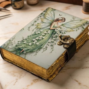 May include: A rectangular journal with gold-edged pages and a metal latch closure. The cover features an illustration of a winged figure in a flowing green dress adorned with white flowers, set against a pale background with vines.