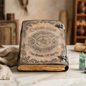 K&ouml;nnte beinhalten: Ein ledergebundenes Buch mit dem Titel "Grimoire The Book Of Spells" mit einem gepr&auml;gten Pentagramm und Mond-Design. Das Buch hat gealterte, vergilbte Seiten und verzierte Metallverschl&uuml;sse. Der Einband ist hellbraun.