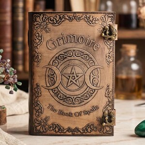 K&ouml;nnte beinhalten: Ein braunes, ledergebundenes Buch mit dem gepr&auml;gten Wort "Grimoire" auf dem Einband. Der Einband zeigt ein Pentagramm in einem Kreis und Halbmonde. Der Text "The Book Of Spells" ist ebenfalls sichtbar. Das Buch hat Metallverschl&uuml;sse.