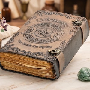 K&ouml;nnte beinhalten: Ein ledergebundenes Buch mit dem Wort "Grimoire" und "The Book Of Spells" auf dem Einband. Der Einband zeigt ein Pentagramm und ein Mondsichel-Design. Das Buch ist mit einem Lederriemen und Metallverschl&uuml;ssen gesichert. Die Seiten sind gealtert.