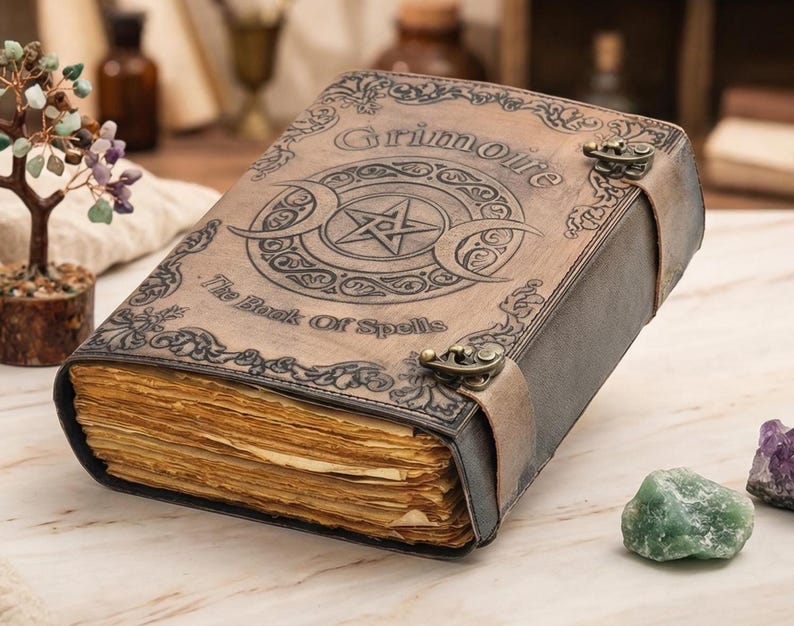 K&ouml;nnte beinhalten: Ein ledergebundenes Buch mit dem Wort "Grimoire" und "The Book Of Spells" auf dem Einband. Der Einband zeigt ein Pentagramm und ein Mondsichel-Design. Das Buch ist mit einem Lederriemen und Metallverschl&uuml;ssen gesichert. Die Seiten sind gealtert.