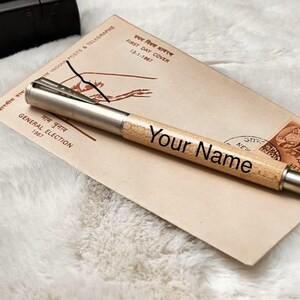 K&ouml;nnte beinhalten: Ein personalisierter Holzstift mit silbernen Akzenten, graviert mit "Your Name". Der Stift liegt auf einem Vintage-Umschlag mit Text in Englisch und einer anderen Sprache sowie einer Briefmarke. Der Hintergrund ist eine weiche, wei&szlig;e, strukturierte Oberfl&auml;che.