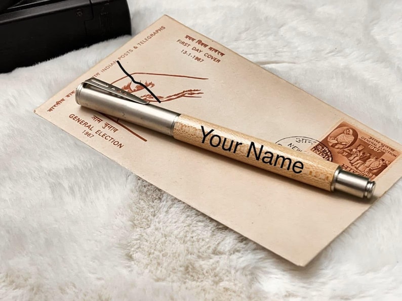 K&ouml;nnte beinhalten: Ein personalisierter Holzstift mit silbernen Akzenten, graviert mit "Your Name". Der Stift liegt auf einem Vintage-Umschlag mit Text in Englisch und einer anderen Sprache sowie einer Briefmarke. Der Hintergrund ist eine weiche, wei&szlig;e, strukturierte Oberfl&auml;che.