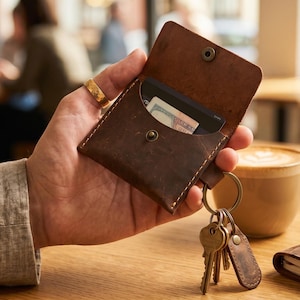 Könnte beinhalten: Ein braunes Lederportemonnaie mit Druckknopfverschluss, das eine Kreditkarte und Bargeld enthält. Das Portemonnaie ist an einem Schlüsselring mit Schlüsseln befestigt. Eine Kaffeetasse und ein kleines Notizbuch sind im Hintergrund.