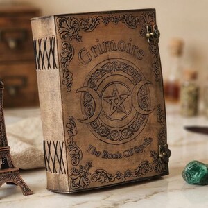 K&ouml;nnte beinhalten: Ein braunes, ledergebundenes Buch mit den Worten "Grimoire" und "The Book Of Spells" auf dem Einband. Der Einband zeigt ein Dreifachmondsymbol mit einem Stern in der Mitte. Das Buch hat Metallverschl&uuml;sse und dekorative N&auml;hte am R&uuml;cken.