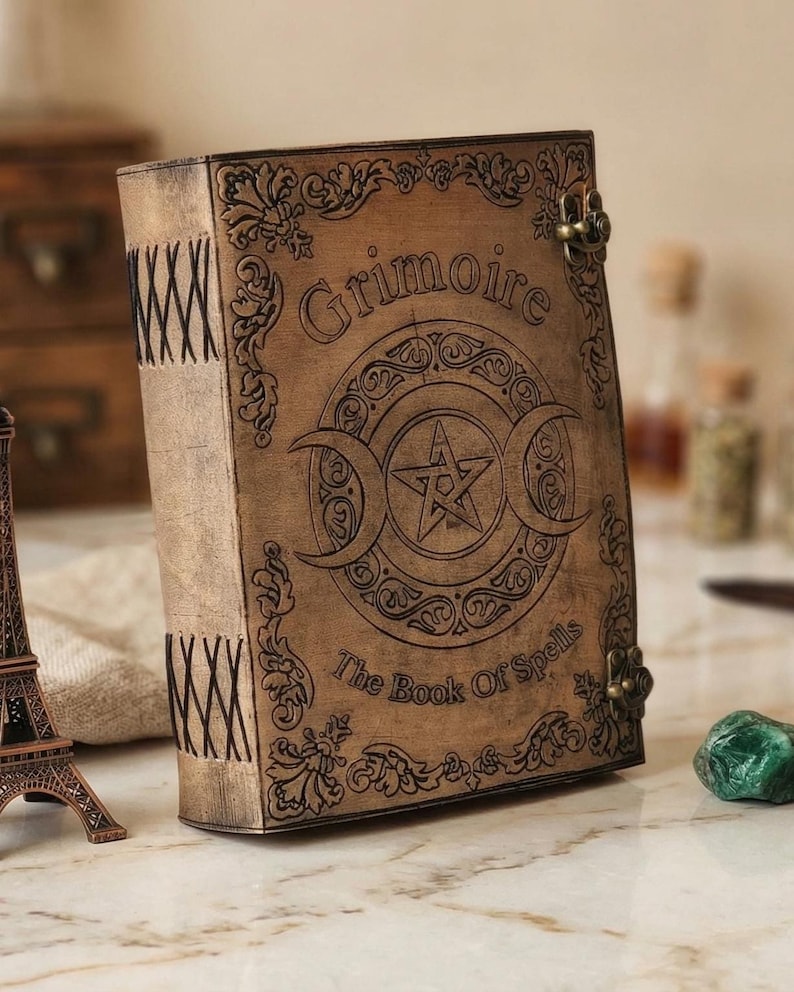 K&ouml;nnte beinhalten: Ein braunes, ledergebundenes Buch mit den Worten "Grimoire" und "The Book Of Spells" auf dem Einband. Der Einband zeigt ein Dreifachmondsymbol mit einem Stern in der Mitte. Das Buch hat Metallverschl&uuml;sse und dekorative N&auml;hte am R&uuml;cken.