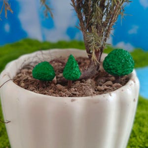 Puede incluir: Una maceta de cerámica blanca con estrías verticales contiene tierra, una pequeña planta leñosa y tres figuras de árboles verdes en miniatura. Las figuras incluyen dos árboles verdes redondeados y un árbol verde cónico, situados en la tierra.