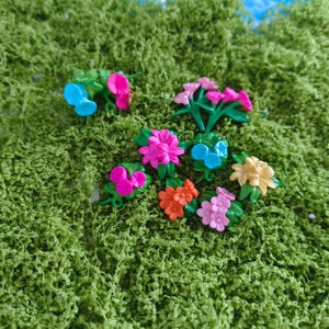 Pode incluir: Uma coleção de pequenas flores de plástico coloridas, em tons de rosa, azul, laranja e amarelo, com folhas verdes. As flores estão dispostas sobre um leito de musgo verde, criando uma cena vibrante e alegre.