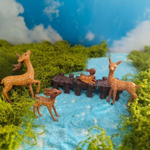 May include: A set of three small deer figurines in a light brown colour with white spots. One deer stands on the left, one stands on the right, and one small fawn rests on a dark brown miniature wooden bridge spanning a blue stream.