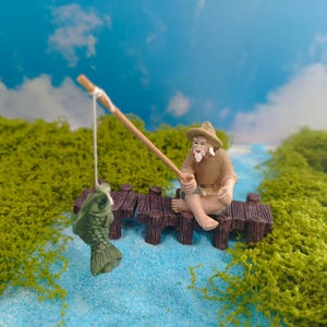 May include: A miniature figurine of an elderly person wearing a tan shirt and a straw hat, sitting on a dark brown wooden pier. The figure holds a thin wooden fishing rod with a white string attached to a small green fish hanging in the air.