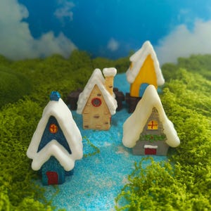 Pode incluir: Quatro casas em miniatura com telhados cobertos de neve dispostas sobre uma superfície azul. As casas apresentam cores variadas, incluindo azul, bege, amarelo e cinza, com pequenas janelas, portas e chaminés. Rodeadas por musgo verde.