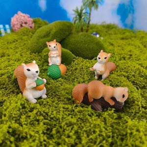 Puede incluir: Cuatro figuras de ardillas en miniatura en varias poses sobre un lecho de musgo verde. Una ardilla sostiene una bellota, otra está sentada encima de una bellota y una tercera descansa sobre un tronco. Las ardillas tienen pelaje marrón y blanco.