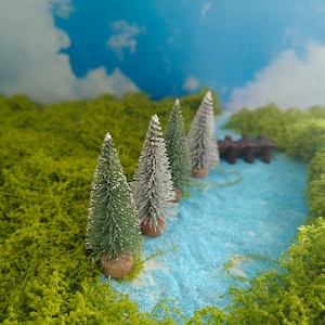 Könnte beinhalten: Eine Miniaturlandschaft mit vier kleinen Flaschenbürstenbäumen mit grünen und silbernen Nadeln, die jeweils auf einem runden Holzsockel stehen. Die Bäume sind entlang eines hellblauen, strukturierten Pfades angeordnet, der von hellgrünem Moos gesäumt ist.