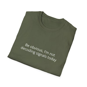 May include: Olive green t-shirt with the text "Be obvious, I'm not decoding signals today" in white. The shirt is folded and appears to be made of a soft material. The neckline is a classic crew neck.