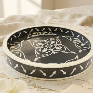 May include: A decorative black and white tray with an intricate floral design sits on a marble surface. A cream-colored notebook and a gold pen are next to the tray. A white orchid flower is also present.