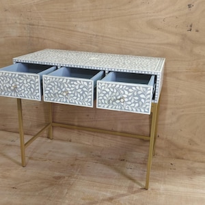 May include: A white and grey floral patterned desk with three drawers, each with a silver knob. The desk has a gold metal frame. The drawers are open, revealing a light blue interior.
