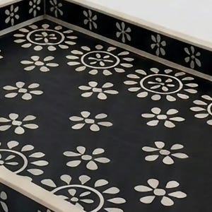 May include: Rectangular serving tray with a black surface adorned with white floral patterns. The tray's border is a light beige color, and the floral design is repeated around the edges. A decorative item for serving or display.