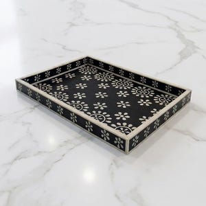 May include: Rectangular black decorative tray with a floral pattern in white. The tray has a white border and is set on a white marble surface. A stylish accent piece for home decor.