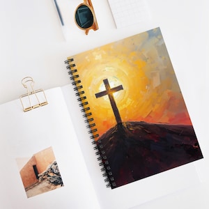 Puede incluir: Cuaderno de espiral con una pintura artística de una cruz sobre una colina contra un fondo amarillo y naranja brillante. El cuaderno tiene una encuadernación en espiral negra y está abierto sobre una superficie blanca.