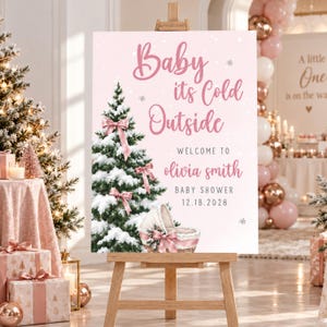 May include: A rectangular baby shower welcome sign on a wooden easel featuring a pink background, a snow-covered Christmas tree with pink bows, and a bassinet. The text reads: Baby its Cold Outside, WELCOME TO olivia smith BABY SHOWER 12.18.2028.