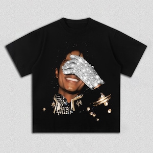 May include: Black t-shirt featuring a portrait of a smiling person with a sparkling glove covering their eyes. The person is wearing a black jacket with gold and silver embellishments. The image has a dark background.
