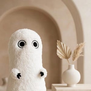 Pode incluir: Um brinquedo de pelúcia em forma de fantasma com tecido branco felpudo, dois grandes olhos circulares pretos com contornos brancos e braços pequenos que terminam em três garras pretas. A criatura tem uma cabeça arredondada.