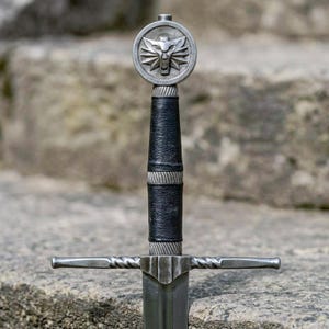 May include: A sword hilt featuring a black leather-wrapped grip with silver-toned metal accents. The pommel is a circular silver-toned disc embossed with a stylized wolf head emblem. The crossguard is a straight, twisted silver-toned metal bar.