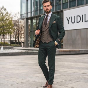 Puede incluir: Un traje de tres piezas verde oscuro con chaleco estampado, camisa blanca y corbata marrón. El traje se combina con zapatos de cuero marrón y un reloj dorado. El fondo presenta un edificio moderno con la palabra "YUDIL" en un letrero.