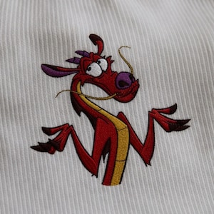 May include: Embroidered red dragon with yellow accents and purple details on a white ribbed fabric. The dragon has an expressive face and is a cartoon character. The design is likely for apparel or home decor.
