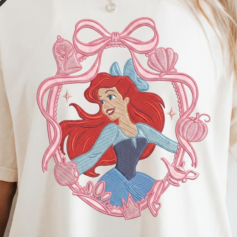 May include: Embroidered design featuring a cartoon character with red hair, wearing a blue dress, and framed by a pink ribbon and various themed elements. The design is on a cream-colored fabric.