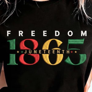 Op de afbeelding: Zwart T-shirt met het woord "FREEDOM" in wit boven de gekleurde cijfers "1865". Het woord "JUNETEENTH" staat onder de cijfers, met sterren aan weerszijden. De cijfers zijn groen, rood en goud.