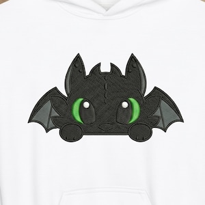Può includere: Un disegno di drago nero ricamato con occhi verdi e ali grigie è presente su una felpa con cappuccio bianca. Il drago ha un'espressione amichevole, con le zampe visibili. Il disegno è centrato sul petto della felpa.