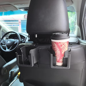 以下が含まれることがあります： 黒い車のシートオーガナイザー。2つのカップホルダーと小さな収納コンパートメントがあります。赤と白のデザインの使い捨てコーヒーカップがカップホルダーの1つに入っています。オーガナイザーは車のシートの背面に装着します。
