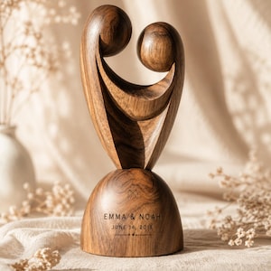 May include: A dark brown wooden sculpture featuring two abstract figures embracing. The base is engraved with the text EMMA & NOAH, JUNE 14, 2019, and a small heart symbol. The wood grain is visible throughout the smooth, curved surface of the piece.