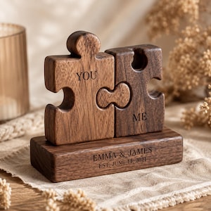 May include: Two interlocking dark brown wooden puzzle pieces mounted on a rectangular wooden base. The left piece is engraved with the word YOU and the right piece with ME. The base is engraved with EMMA & JAMES and EST. JUNE 14, 2021.