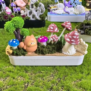 May include: A miniature diorama in a white rectangular tray featuring a small orange squirrel figurine, a green tree, and several mushrooms in red, blue, and purple. The scene includes green moss, a small beige fence, and a sandy base texture.