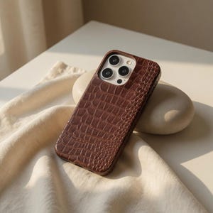 Pode incluir: Uma capa de telefone castanha com textura de pele de crocodilo. A capa tem formato retangular com cantos arredondados e um recorte para a câmera. A capa está sobre uma superfície branca com um tecido bege.