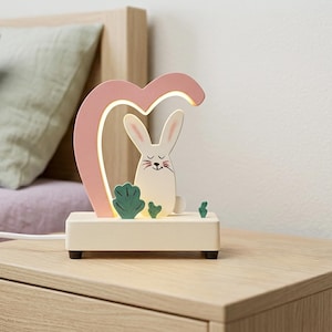 Op de afbeelding: Decoratieve nachtlamp met een wit konijn in een roze hartvormige lijst. Het konijn rust op een crèmekleurige basis met groene bladeren. De lamp staat op een houten nachtkastje.
