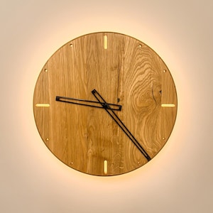 Könnte beinhalten: Eine kreisförmige Wanduhr aus hellbraunem Holz mit sichtbarer natürlicher Maserung. Die Uhr verfügt über dünne schwarze Metallzeiger und beleuchtete Schlitze an den Stundenpositionen, die einen sanften Schein am Rand erzeugen.
