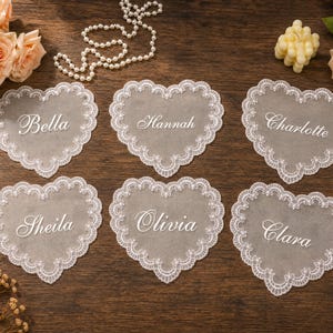 May include: Six heart-shaped white lace doilies featuring scalloped edges and embroidered names Bella, Hannah, Charlotte, Sheila, Olivia, and Clara in white script font, arranged on a dark wooden surface with peach roses and pearl beads.