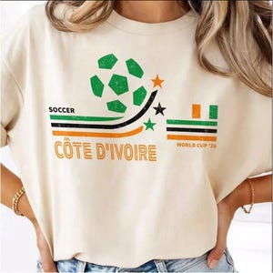 May include: A cream-colored t-shirt featuring a graphic with a green soccer ball, three stars, and horizontal stripes in green, black, and orange. The text reads SOCCER, COTE D'IVOIRE, and WORLD CUP '26 in a distressed, vintage-style font.