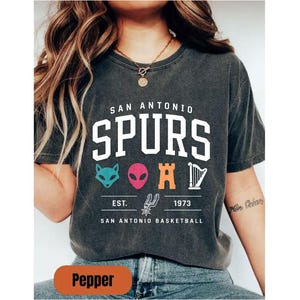 May include: Dark grey t-shirt featuring the text "SAN ANTONIO SPURS" in white. Below are four colourful graphics and the text "EST. 1973 SAN ANTONIO BASKETBALL". The shirt is a unisex style.