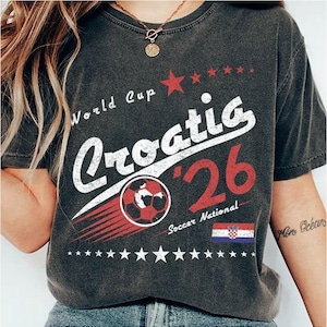 May include: A charcoal gray t-shirt featuring a distressed graphic print that reads World Cup Croatia '26 Soccer National. The design includes a red soccer ball, a row of stars, and the Croatian flag in red, white, and blue colors.