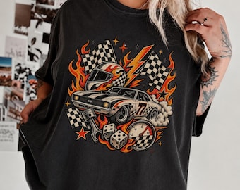 Puede incluir: Una camiseta gris carbón con un estampado gráfico de un coche deportivo blanco con el número 77, un casco de carreras, banderas a cuadros, un pistón, dados, un velocímetro y llamas naranjas, estrellas y rayos.