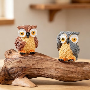 May include: Two small owl figurines perched on a piece of driftwood. The left owl is brown with white facial discs and orange eyes. The right owl is gray with white facial discs and orange eyes. Both have textured feathers and orange feet.