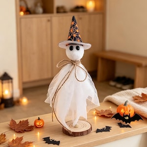 Op de afbeelding: Een wit spookfiguurtje met zwarte ogen, met een zwarte heksenhoed met een oranje spinnenwebpatroon. Het spook heeft een lichaam van doorschijnende witte tule, vastgebonden met een bruin touwstrikje, op een ronde houten voet met jaarringen.