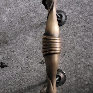 May include: A vertical cabinet handle with a brushed antique brass finish. The handle features a central section with horizontal ribbed detailing, flanked by smooth, tapered segments and rounded finials at both ends, secured by two circular mounts.