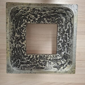 May include: Square, textured, metallic-looking decorative tray with a central square opening. The tray has a rough, mottled surface in shades of gray and bronze, with a lighter-colored interior.